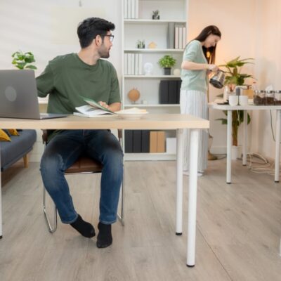Mężczyzna siedzi przy biurku z regulowanym stelażem, podczas gdy kobieta przygotowuje napoje w tle.