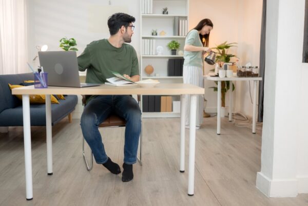 Mężczyzna siedzi przy biurku z regulowanym stelażem, podczas gdy kobieta przygotowuje napoje w tle.